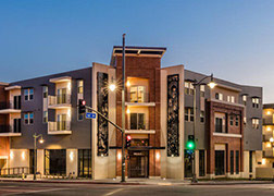 Evening view with lighting: artwork frames the corner entrance of contemporary style three-story apartment building clad in stucco, red brick, and gray siding.